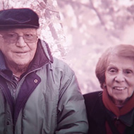 Um homem idoso à esquerda, de boina, óculos e casaco verde, à direita uma mulher idosa de casaco preto e gola vermelha, ambos sorrindo.